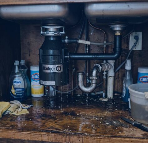 A leaking garbage disposal under a kitchen sink with standing water on the cabinet floor, demonstrating the mess caused when learning how to unclog a garbage disposal with standing water.