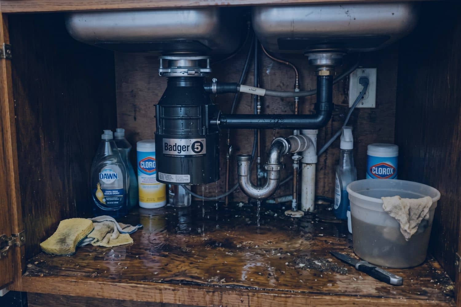 A leaking garbage disposal under a kitchen sink with standing water on the cabinet floor, demonstrating the mess caused when learning how to unclog a garbage disposal with standing water.