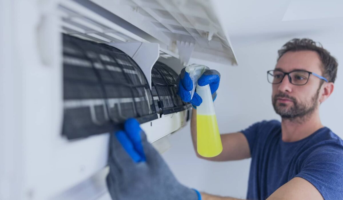 A man in work gloves sprays a yellow cleaning solution onto the internal filters of a wall-mounted air conditioning unit to perform maintenance.
