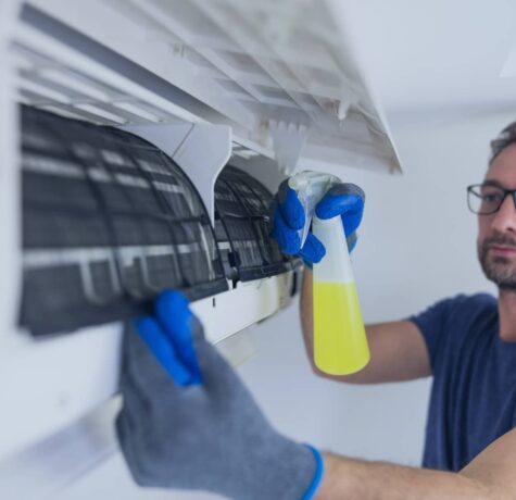 A man in work gloves sprays a yellow cleaning solution onto the internal filters of a wall-mounted air conditioning unit to perform maintenance.