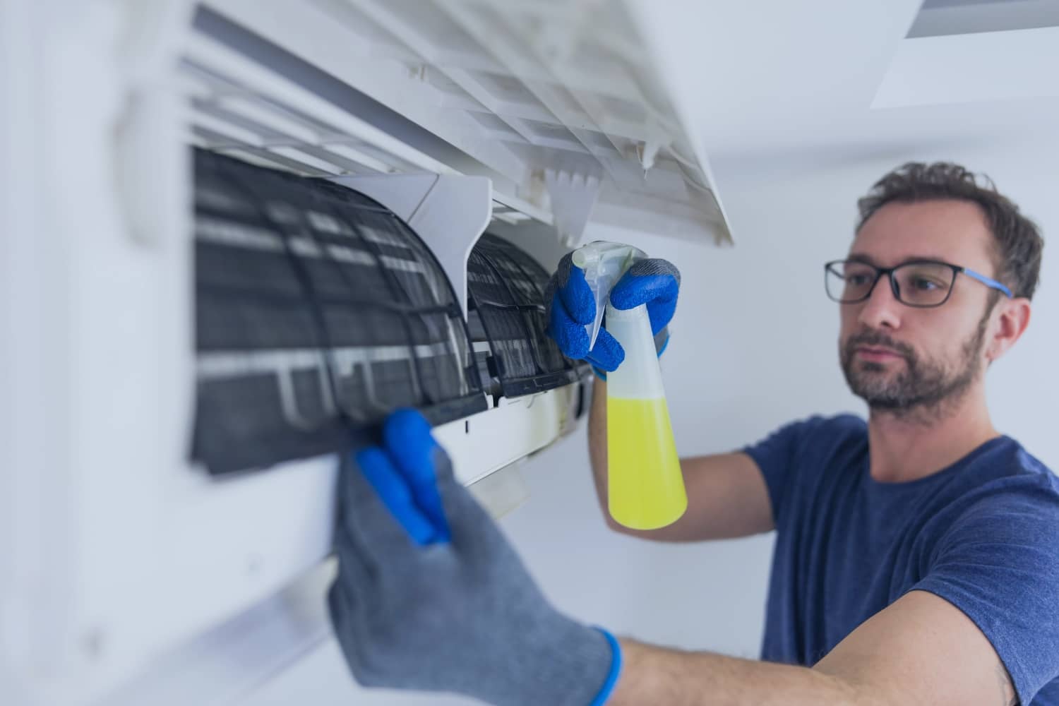 A man in work gloves sprays a yellow cleaning solution onto the internal filters of a wall-mounted air conditioning unit to perform maintenance.