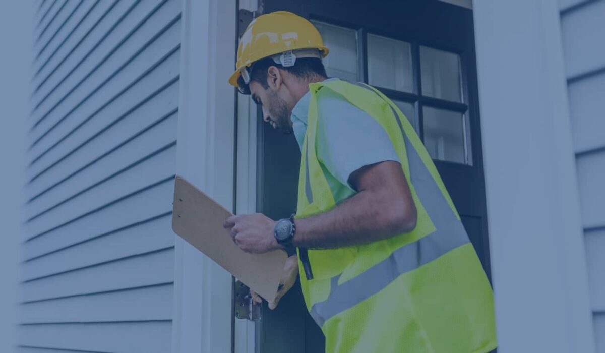 A home inspector wearing a hard hat and high-visibility vest holds a clipboard while inspecting a residential door frame.