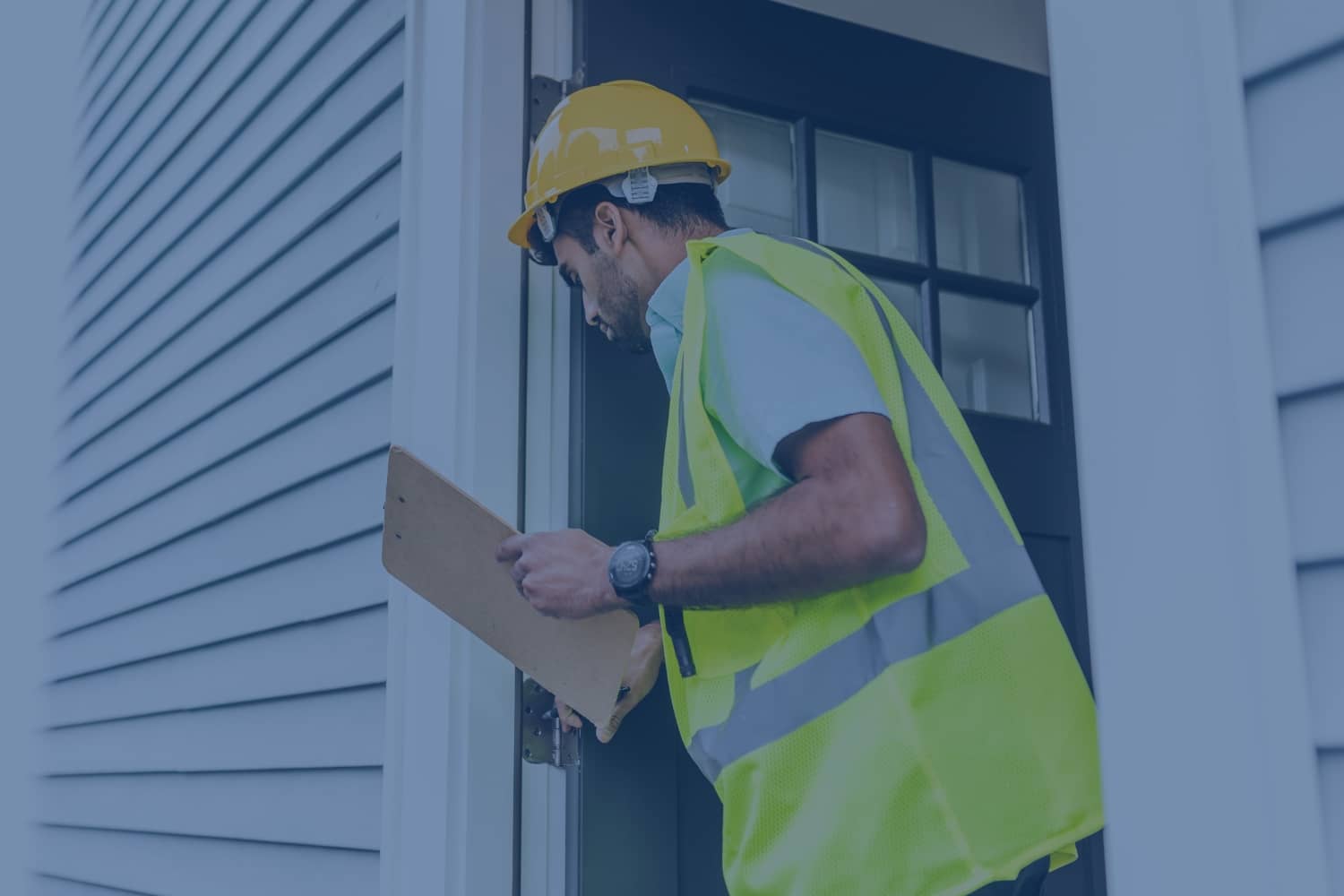 A home inspector wearing a hard hat and high-visibility vest holds a clipboard while inspecting a residential door frame.