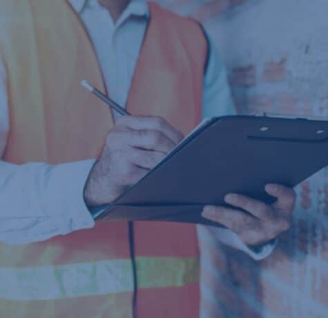 A commercial real estate inspector wearing a high-visibility safety vest writes notes on a clipboard while evaluating a building wall