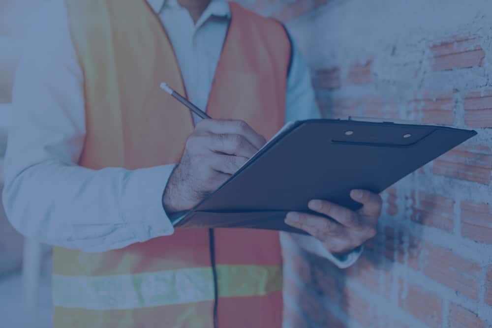 A commercial real estate inspector wearing a high-visibility safety vest writes notes on a clipboard while evaluating a building wall
