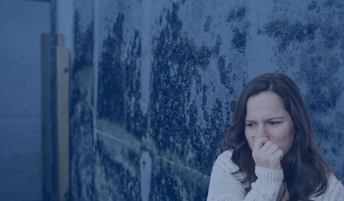 A woman in a white sweater covers her nose with her hand in discomfort while standing next to a wall covered in extensive dark mold.