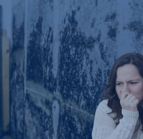 A woman in a white sweater covers her nose with her hand in discomfort while standing next to a wall covered in extensive dark mold.