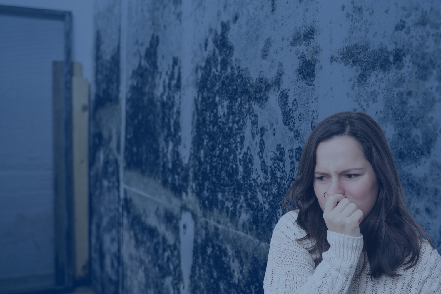A woman in a white sweater covers her nose with her hand in discomfort while standing next to a wall covered in extensive dark mold.